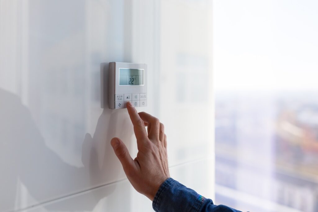 A person's hand adjusting a modern digital thermostat mounted on a white wall, setting the temperature to 22 degrees.
