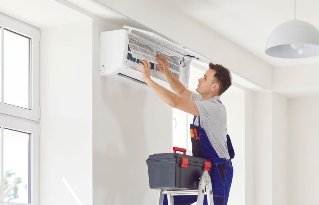 new hvac system cost Male worker from AC maintenance service, standing on ladder with toolbox, opens modern white air conditioner on wall, repairs some inside parts and changes filters